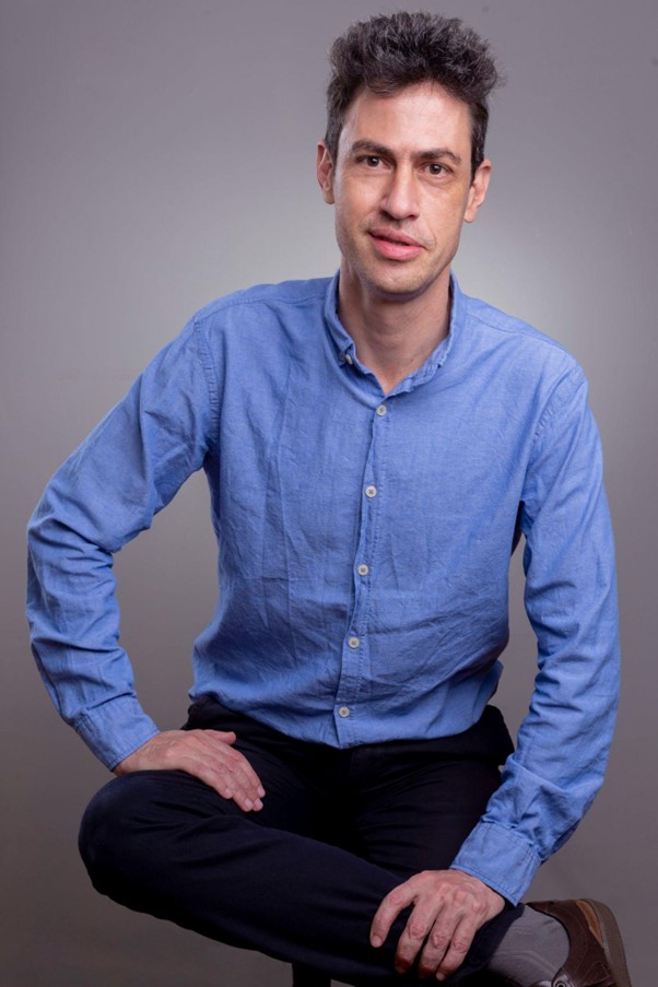 Foto de Mário Mello sentado e sorrindo. Ele é um homem branco de cabelo castanho escuro curto, usa uma camisa azul e uma calça azul-marinho. Suas mãos estão apoiadas na perna direita que está cruzada sob a esquerda.